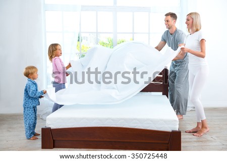 Photo of loving family of four making bed. Young family demonstrating quality of mattress and holding blanket