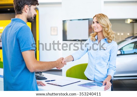 Photo of young female consultant and buyer. Young man buying new car in car showroom. Signing of contract and insurance. Man and woman shaking hands