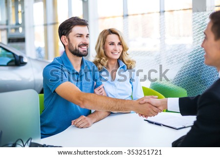 Photo of young male consultant and couple. Young woman and man buying new car in auto show. Signing of contract and shaking hands. Concept for car rental