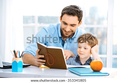 Nice family photo of little boy and his father. Boy and dad sitting at room with big window. Young man helping his son to make homework