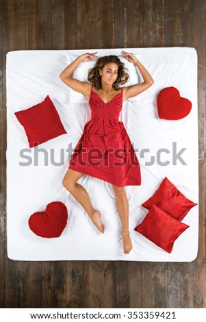 Top view photo of beautiful mixed-race girl sleeping on big white bed. Young pretty woman wearing nice red dress. Girl lying on back. There are red pillows around her
