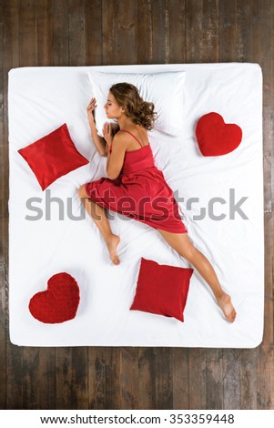 Top view photo of beautiful mixed-race girl sleeping on big white bed. Young pretty woman wearing nice red dress. Girl lying on stomach. There are red pillows around her