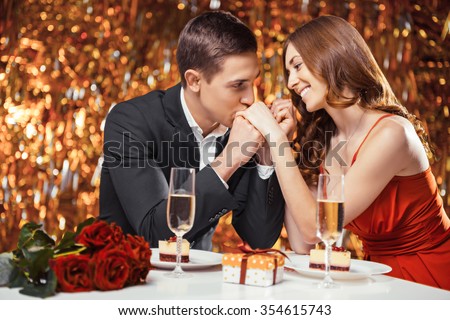Romantic photo of beautiful couple on glitter gold background. Couple having date at Valentine\'s Day. Lovers having dinner. There are glasses with champagne, desserts, roses and gift on table