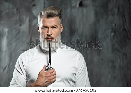 Portrait of stylish professional hairdresser with beard. Man wearing shirt, looking at camera and holding scissors near his beard