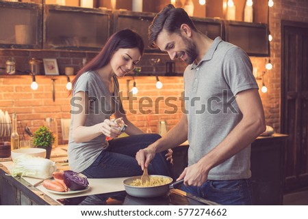 Young beautiful couple in kitchen. Family of two preparing food. Woman sitting on kitchen tabletop and helping man to make delicious pasta. Nice loft interior with light bulbs