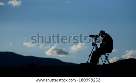 Panoramic Silhouette of Big Game Hunter / Wolf Watcher / BIrd Watcher using spotting scope telescope optics to view distant wildlife from a folding chair, with blue sky and clouds in the background