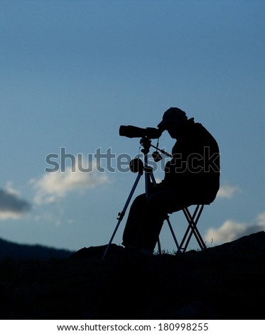 Silhouette of Big Game Hunter / Wolf Watcher / BIrd Watcher using spotting scope telescope optics to view distant wildlife from a folding chair, with blue sky and clouds in the background