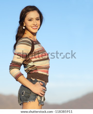 Pretty young woman wearing stylish cut denim shorts and sweater with blue sky and desert mountains in background