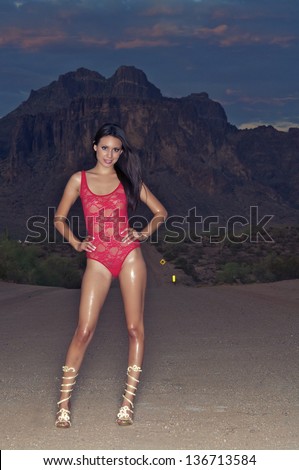 Sexy beautiful young woman standing posing outdoors in red one-piece lingerie with mountain desert background.