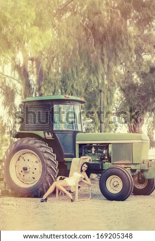 Fashion style photo of young woman sitting in chair beside farm tractor machinery.  Sun light rays filtering through trees.