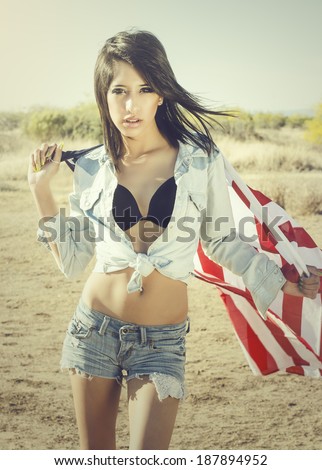 Beautiful young woman wearing denim shorts and top holding American flag in desert location