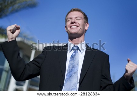 Happy smiling triumphant business man with hands and arm raised to air standing outside corporate head corporate office.