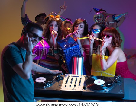 semi-profile of dj mixing music, with a goup of girls blowing party horns to him, holding presents, and  other people dancing in the background