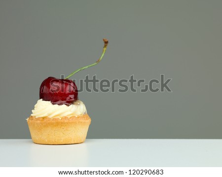 small tart topped with vanilla cream and a cherry, on white table and neutral background