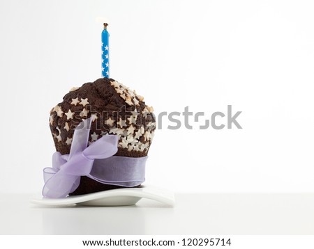 single chocolate muffin on white plate, decorated with white sugar stars, wrapped in purple ribbon and bow, with blue candle on top, on white background