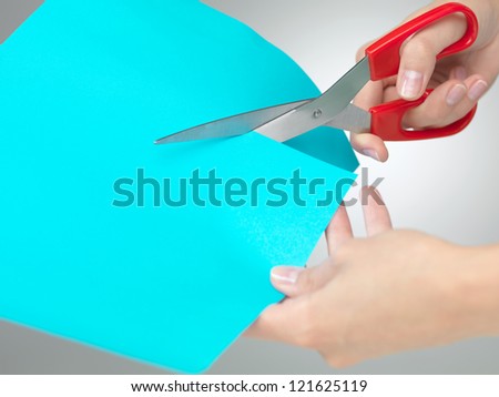 hands cutting a shape from a blue paper with a pair  of red scissors on neutral grey background