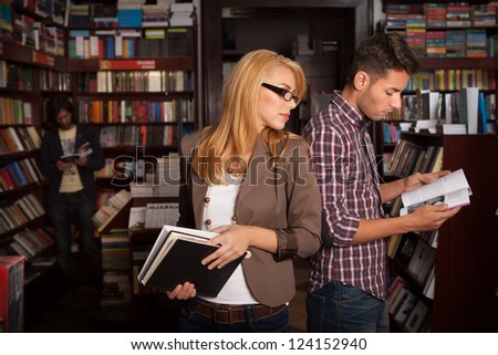 two caucasian young people standing back to back in a bookshop with books in their hands with other bookshelves in the background