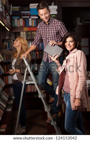 young caucasian guy up on a ladder in a library taking a book from an attractive girl, with another young girl in background choosing a book from a shelf
