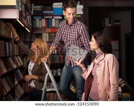 young caucasian guy up on a ladder in a library taking a book from an attractive girl, with another young girl in background choosing a book from a shelf