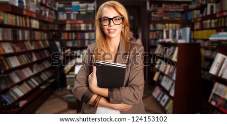 close-up of young attractive caucasian girl with geeky eyeglasses holding two books in her arms with many bookshelves in background