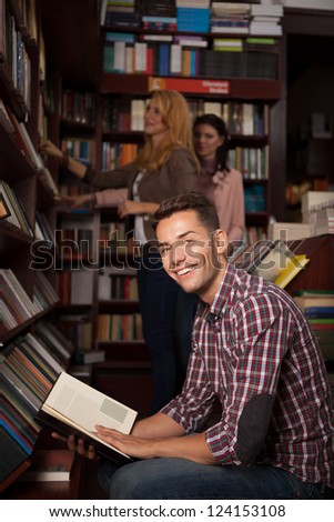 young caucasian handsome guy in a bookstore smiling with an opened book in his hands, with other people in background