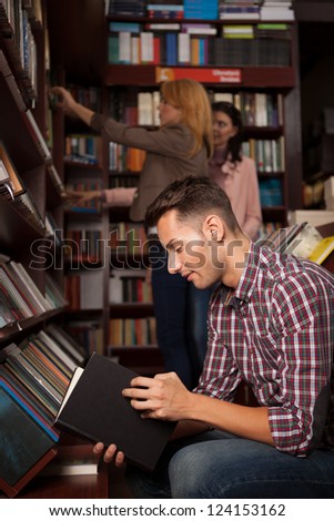 young caucasian handsome guy in a bookshop reading a book with other people in background