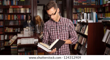 close-up of young caucasian man with eyeglasses in a bookstore with an opened book in his hands reading something with other bookshelves in background
