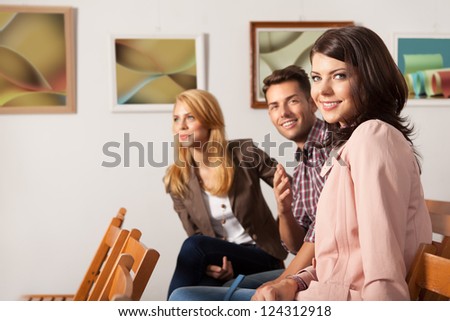 close-up of three attractive young people smiling and sitting down on wooden chairs with framed abstract photography works on background
