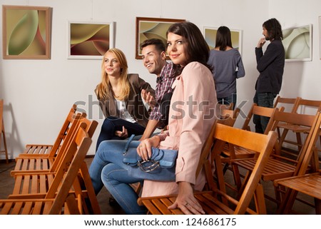 young attractive people sitting down on wooden chairs at an abstract photography exhibition, enjoying themselves, with other people looking at art works on background