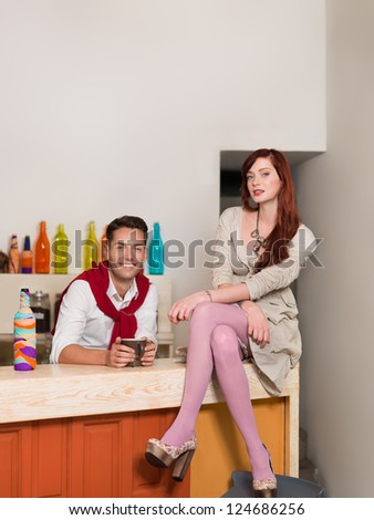 young attractive red haired caucasian girl sitting on the counter with legs and arms crossed with a handsome guy behind the counter in colorful cafe