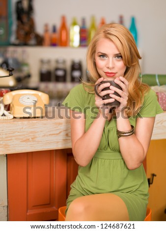 young attractive blonde girl sitting on a chair near the conter of a colorful cafe with a coffee mug in her hands