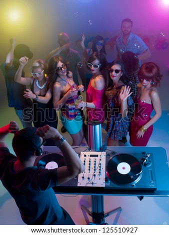 upper view of dj standing and playing music with turntables at a party with colorful lights and happy young people dancing with sunglasses and soap bubbles