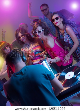 dj mixing music with turntables and headphones in front of young dancing people with sunglasses and cocktails surounded by colorful lights