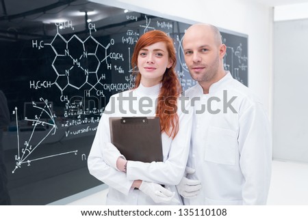 general-view of a teacher and a student in a chemistry lab smiling in the camera and a blackboard on the background