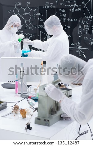 close-up of three people in a laboratory testing and applying chemical procedures using transparent tools and  colorful liquids on a lab table with a blackboard with formulas on the background