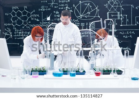 general-view of one man in a chemistry lab supervising two women manipulating lab tools with colorful liquids and seedlings on a lab table with a blackboard on the background
