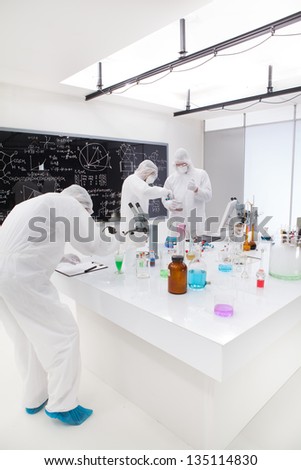 general view  of people working in a chemistry lab around a lab table with colorful liquids and lab tools with a blackboard on the background.