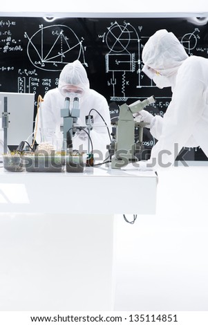 general-view of two scientists studying in a chemistry lab manipulating microscopes on a lab worktable with a blackboard on the background