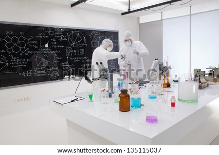 general view of two people making a laboratory experiment around a laboratory table with laboratory tools with colorful liquids and a blackboard with chemical formulas on the background