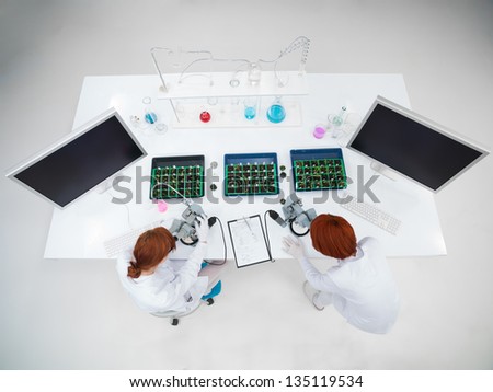 bird-eye of two women in a chemistry lab analysing under microscope on a lab table with with seedling boxes  and two pc monitors