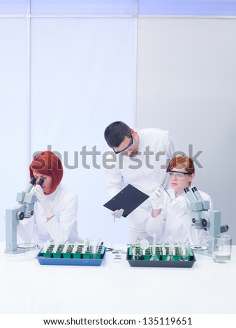 general view  of a teacher in a chemistry lab supervising the activity on two students analyzing plants on a lab table and  observing reactions under microscope
