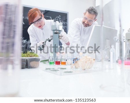 close-up of a student in a chemistry lab around lab tools and colorful liquids conducting an experiment under microscope under the observation of her teacher