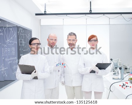 general-view of four co-workers in a chemistry lab around lab table with lab tools and colorful liquids and a blackboard on the background