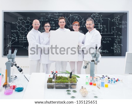 close-up of five smiling  scientists in a chemistry lab holding in hands an empty banner and confident looking in the camera with a blackboard on the background