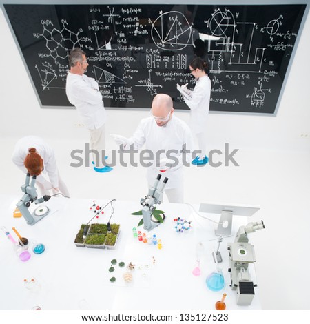bird-eye of four people in a chemistry lab conducting experiments , analyzing substances around a lab table and writing on a blackboard