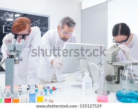 general-view of two students in a chemistry lab analyzing under microscope around lab tools and colorful liquids while the teacher is looking on a pc monitor