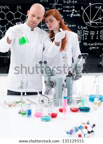 general-view of a teacher conducting a lab experiment and a student  watching the procedure around the lab table with lab tools and a blackboard on the background