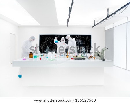 general-view of three scientists in a chemistry lab conducting an experiment around a lab table with colorful liquids and lab tools with a blackboard on the background