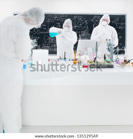 close-up of three scientists in a chemistry lab conducting an experiment around a lab table with colorful liquids and lab tools with a blackboard on the background