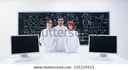general-view of two beautiful girls students and a teacher in a chemistry lab smiling and  holding in hands an empty banner around a lab table with pc monitors and a blackboard on the background